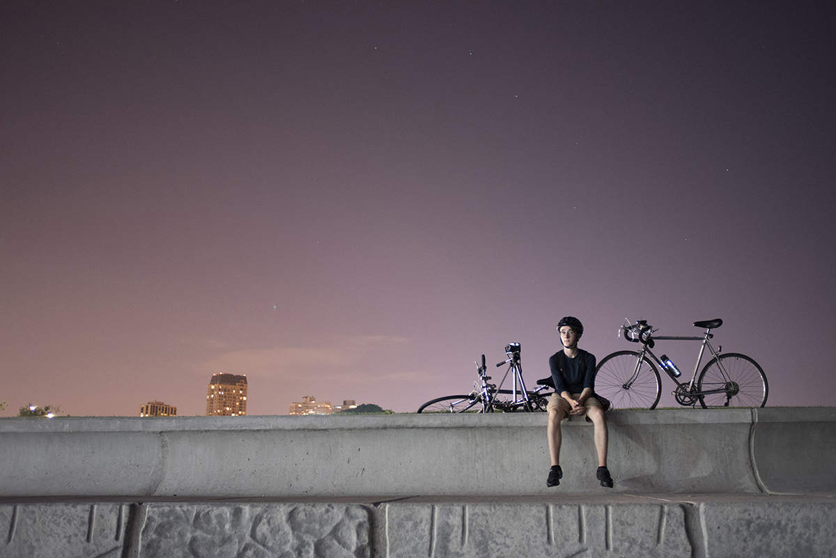 On an incredibly clear summer night in Chicago, my younger brother sits on a ledge next to Lake Michigan. The Big Dipper constellation is barely visible directly above him through the light polluted sky in July 2016.